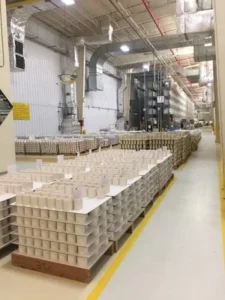 Pallets of stacked material beside an aisle of metal shelving on a manufacturing floor